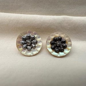 May include: Two round, mother-of-pearl buttons with a central cluster of dark, metallic beads. The buttons have a scalloped edge and are set against a neutral fabric background. The beads have a faceted appearance.