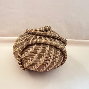 May include: A handwoven basket with a rounded shape, crafted from natural brown and light tan fibers. The basket features a detailed weave pattern and a coiled rim, showcasing a rustic aesthetic.
