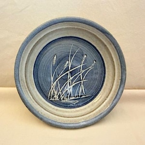 May include: A round ceramic bowl with a blue and beige rim. The center of the bowl features a blue design with white cattails. The bowl is a decorative piece.