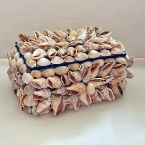May include: A square decorative box covered in various seashells. The box is primarily beige and tan, with a dark blue border. The seashells are of different sizes and shapes, creating a textured surface. The box is likely used for storage or decoration.