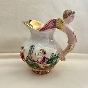 May include: A white ceramic pitcher with a gold rim and a handle shaped like a winged cherub. The pitcher is decorated with a scene of cherubs playing in a garden.