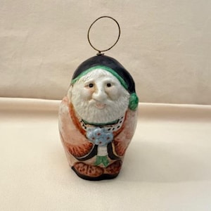 May include: A ceramic figurine of a bearded man with a black hat and green trim. The figurine has a white face, a long white beard, and is holding a small bouquet of blue flowers. A wire loop is attached to the top.