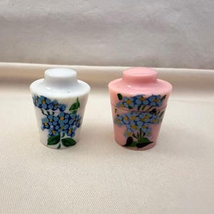 May include: Two small, decorative jars with lids. One is white with blue floral designs and green leaves. The other is pink with similar blue floral designs. Both jars have a cylindrical shape.