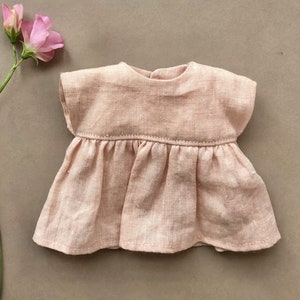 Pode incluir: Um vestido de linho rosa claro com saia franzida e mangas curtas. O vestido &eacute; feito para uma boneca.