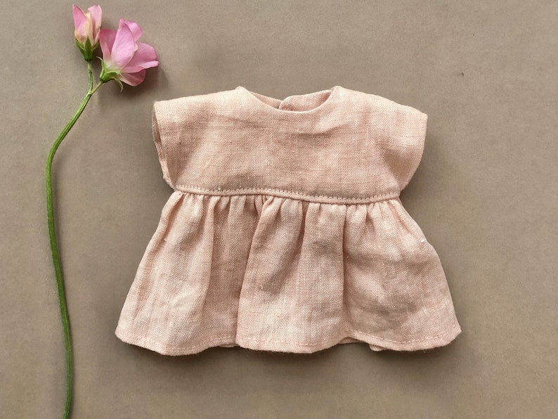Pode incluir: Um vestido de linho rosa claro com saia franzida e mangas curtas. O vestido &eacute; feito para uma boneca.