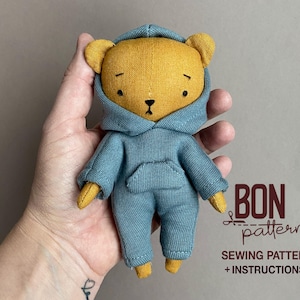May include: A handmade plush bear doll with a yellow head and ears, wearing a blue hooded jumpsuit. The doll has a stitched face and is holding a sewing pattern. The text "BON patterns" and "SEWING PATTERN + INSTRUCTIONS" are visible.