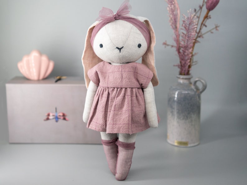 Pode incluir: Uma boneca de coelho de pel&uacute;cia feita &agrave; m&atilde;o com um vestido xadrez rosa e meias rosas. A boneca tem um rosto branco com olhos pretos e um nariz preto. A boneca est&aacute; em p&eacute; sobre uma superf&iacute;cie cinza.