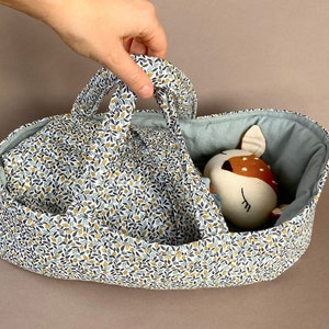 Könnte beinhalten: Eine Stoffpuppen-Tragetasche mit einem Blumenmuster in Blau, Gelb und Weiß. Die Innenseite ist einfarbig hellblau. Ein Stofftier in Form eines Hirsches mit braunen Flecken und einem cremefarbenen Gesicht liegt darin.