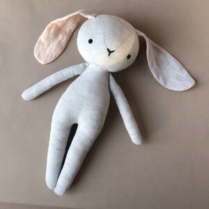 Pode incluir: Um brinquedo de coelho de linho cinza macio com orelhas compridas. Uma orelha &eacute; de cor rosa claro. O coelho tem olhos pretos e um nariz branco.