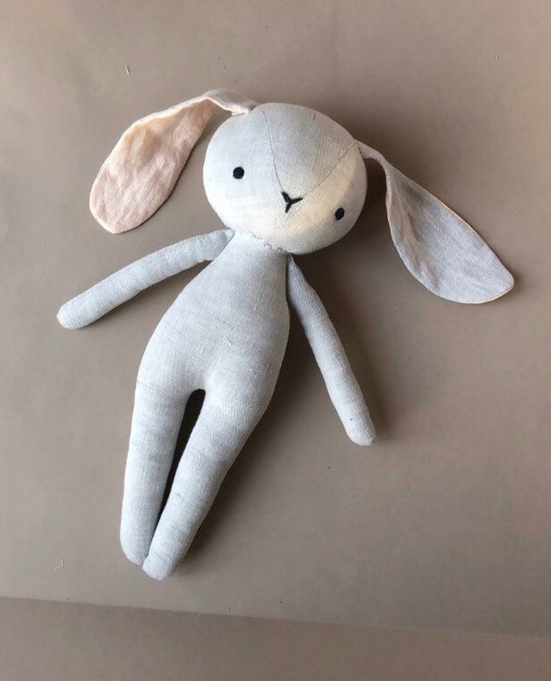 Pode incluir: Um brinquedo de coelho de linho cinza macio com orelhas compridas. Uma orelha &eacute; de cor rosa claro. O coelho tem olhos pretos e um nariz branco.