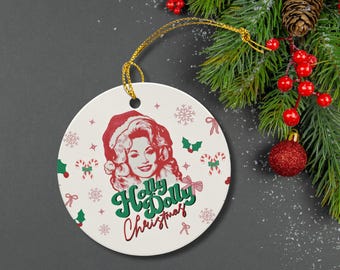 May include: A round white ceramic Christmas ornament featuring a red-toned illustration of a woman wearing a Santa hat. The text "Holly Dolly Christmas" is printed in green and red script. It hangs from a gold string against a dark background.