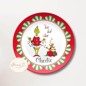May include: A round white plate with a red scalloped border featuring green and white stripes. The center displays a green character draped in colorful Christmas lights, a reindeer in a red gift box, the text ho ho ho, and the name Charlie.