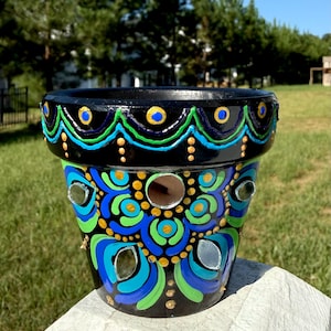 May include: A black ceramic planter featuring a hand-painted mandala design in vibrant shades of blue, green, and gold. The exterior is decorated with small, circular inset mirrors and a scalloped pattern around the rim, creating a detailed texture.