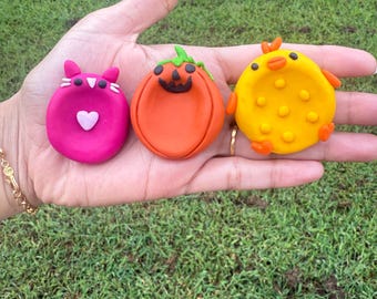 May include: Three colorful clay figures: a pink cat with a heart, an orange pumpkin with a carved face, and a yellow chick with orange accents. The figures are held in a hand against a green grass background.