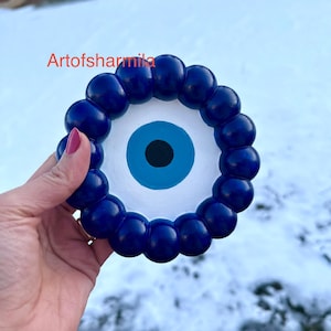 May include: A blue and white decorative eye-shaped object. The eye has a blue iris with a black pupil, surrounded by a ring of blue spheres. The text "Artofsharmila" is visible in the image.