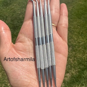 May include: Five silver metal sculpting tools with rounded tips. The tools have textured handles and are held in a hand.