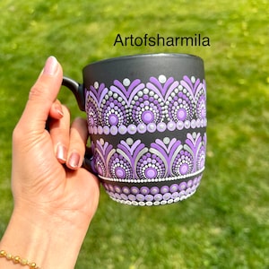 May include: Black ceramic mug with a hand-painted mandala design in shades of purple, silver, and white. The mug features a repeating pattern of dots and floral shapes. The text "Artofsharmila" is visible on the mug.