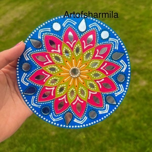 May include: A round, blue, white, pink, green, and yellow mandala with mirrored accents. The mandala is painted on a wooden surface and has a white border.