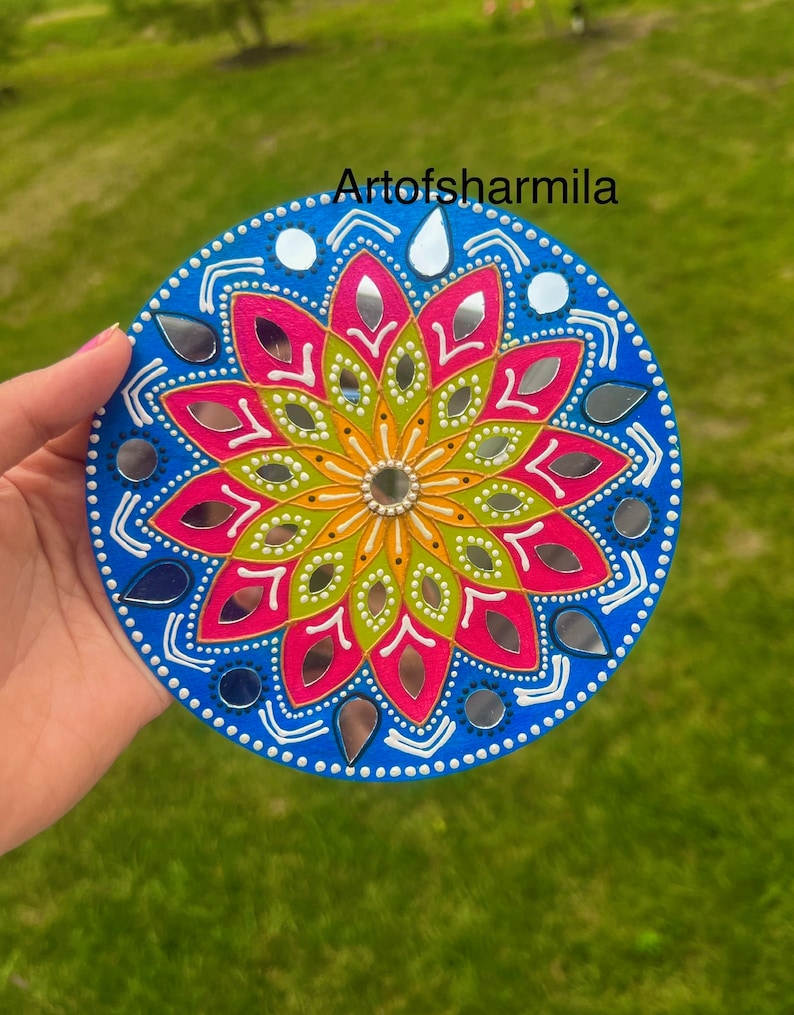 May include: A round, blue, white, pink, green, and yellow mandala with mirrored accents. The mandala is painted on a wooden surface and has a white border.