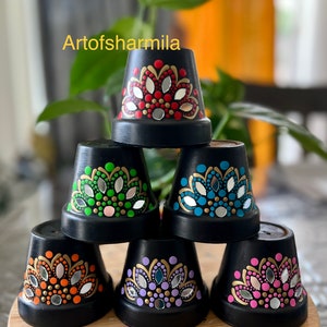 May include: Six black terracotta flower pots with different colored mandala designs painted on them. The designs include red, green, blue, orange, purple, and pink. Each design has a silver mirror accent.