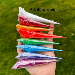 May include: A hand holding a stack of seven colorful tubes of henna paste. The tubes are arranged in a rainbow order, starting with white and ending with purple. The text "Artofsarmilla" is visible at the top of the image.