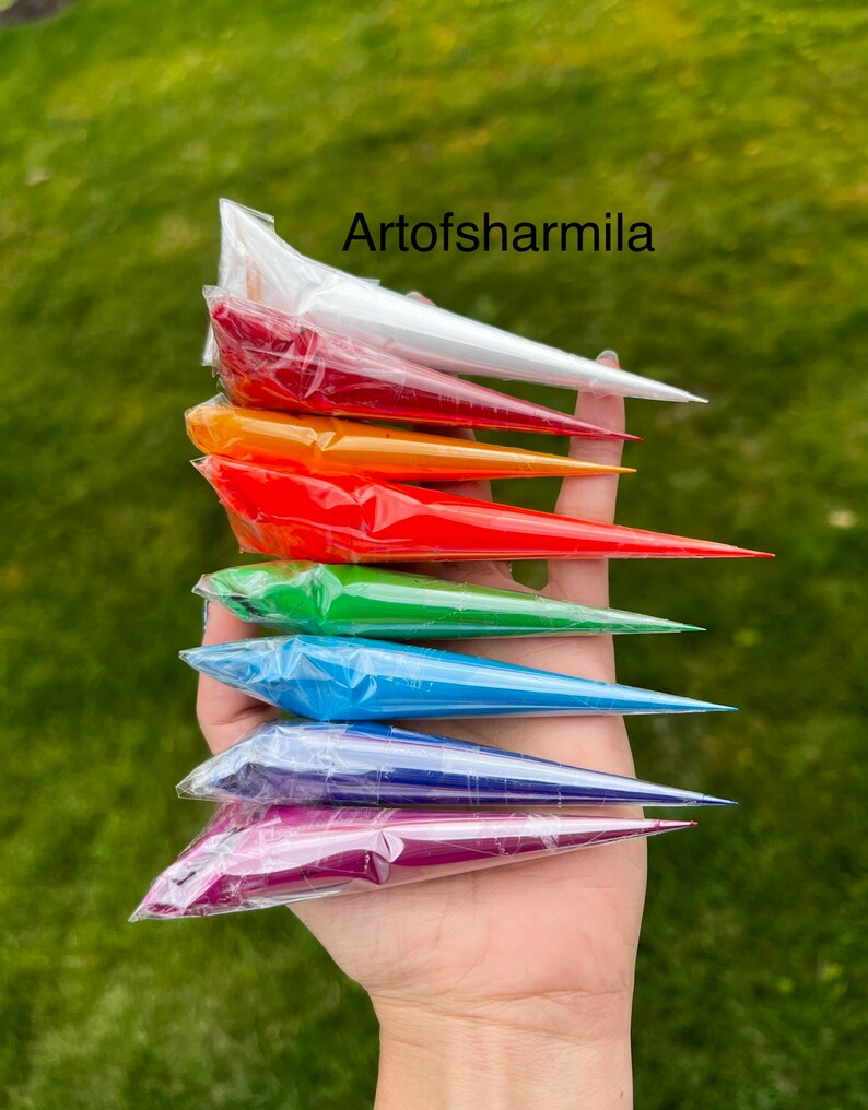 May include: A hand holding a stack of seven colorful tubes of henna paste. The tubes are arranged in a rainbow order, starting with white and ending with purple. The text "Artofsarmilla" is visible at the top of the image.