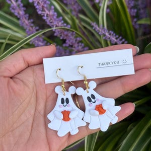 May include: A pair of white ghost earrings with gold-colored hooks. Each earring features a smiling ghost holding an orange pumpkin. The earrings are displayed with a "THANK YOU" card.
