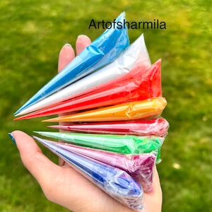 May include: A hand holding a set of eight paint tubes, each with a different color: blue, white, red, orange, yellow, green, purple, and dark blue. The tubes are arranged in a fan shape, with the blue tube at the top and the dark blue tube at the bottom. The tubes are all sealed with plastic wrap.