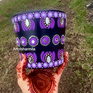 May include: A black painted flower pot with purple floral and mirror accents. The pot has a cylindrical shape and is decorated with repeating patterns of purple flowers, white dots, and small mirrors. The text "Artofsharmila" is visible on the pot.