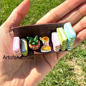 May include: A miniature dark brown wooden shelf featuring five colorful books with gold spine details, a small green potted plant in a white-spotted orange pot, and a tiny cup with two cookies on a plate. The text Artofsharmila is visible.