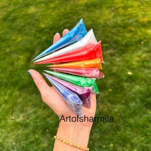 May include: A hand holding a set of eight henna cones in various colors, including blue, white, red, orange, yellow, green, purple, and dark blue. The cones are arranged in a fan shape, with the tips pointing towards the top of the image. The hand is wearing a gold bracelet. The text "Artofsarmilla" is visible in the lower right corner of the image.