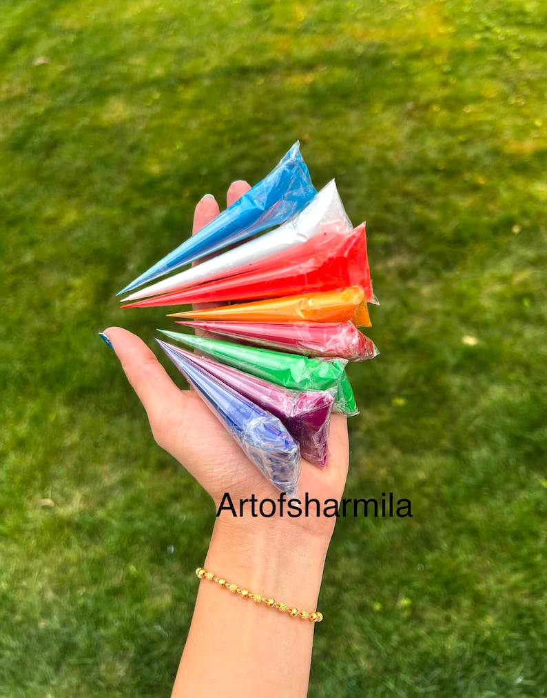 May include: A hand holding a set of eight henna cones in various colors, including blue, white, red, orange, yellow, green, purple, and dark blue. The cones are arranged in a fan shape, with the tips pointing towards the top of the image. The hand is wearing a gold bracelet. The text "Artofsarmilla" is visible in the lower right corner of the image.
