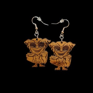 May include: A pair of wooden earrings featuring a cartoon character wearing sunglasses. The character is depicted with a mischievous grin and is holding a small object in each hand. The earrings are a light brown color and have silver hooks.