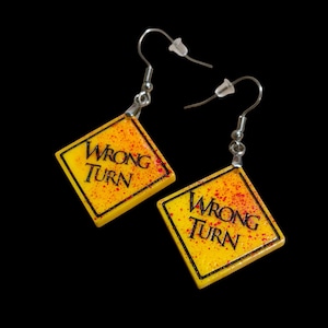 May include: A pair of dangle earrings featuring diamond-shaped yellow charms with black borders and the text WRONG TURN printed in black. The yellow surface is speckled with red paint splatters, and each charm hangs from a silver-toned metal hook.