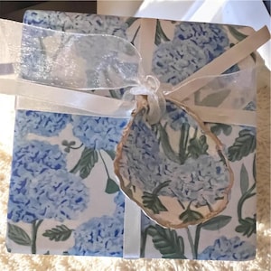May include: A white gift box with a blue and green floral pattern. The box is wrapped with a white ribbon and a sheer white ribbon. A gold-colored shell is attached to the ribbon.