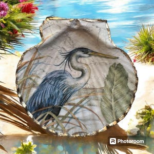 May include: A decorative seashell with a hand-painted image of a blue heron in a marsh setting. The shell has a gold-colored trim and is set against a backdrop of water and foliage. The heron is depicted with detailed feathers and a long beak.
