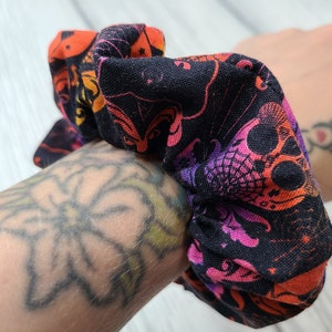 May include: A fabric scrunchie featuring a black background with a repeating pattern of skulls in vibrant shades of orange, pink, and purple. The design includes intricate spiderweb details and stylized facial features printed across the soft material.