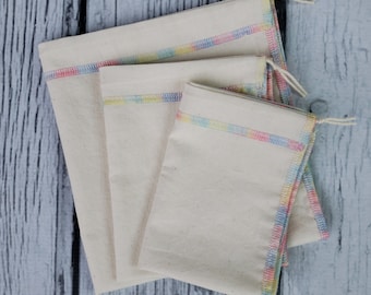 May include: Three white cotton reusable bags with colorful trim. The bags are stacked on top of each other, with the largest bag on the bottom and the smallest bag on top.