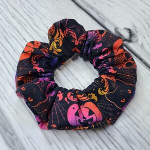 May include: A fabric hair scrunchie featuring a black background with a vibrant, multi-colored Halloween-themed print. The pattern includes orange and pink jack-o'-lanterns, purple spider webs, and swirling decorative motifs throughout the fabric.