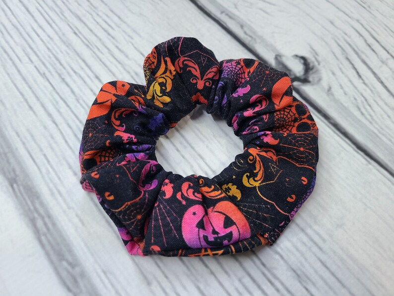 May include: A fabric hair scrunchie featuring a black background with a vibrant, multi-colored Halloween-themed print. The pattern includes orange and pink jack-o'-lanterns, purple spider webs, and swirling decorative motifs throughout the fabric.