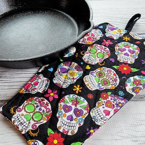 Puede incluir: Un guante de cocina negro con un colorido patrón de calaveras de azúcar, con calaveras blancas con diseños florales y decorativos. El guante está junto a una sartén de hierro fundido negra. El guante tiene un lazo negro para colgar.