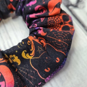 May include: A black fabric hair scrunchie featuring a vibrant Halloween-themed print. The pattern includes orange and pink jack-o'-lanterns, intricate spider webs, and purple accents scattered across the dark background in a repeating design.