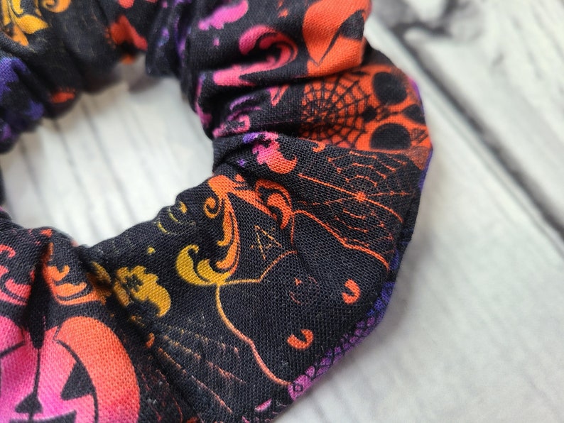 May include: A black fabric hair scrunchie featuring a vibrant Halloween-themed print. The pattern includes orange and pink jack-o'-lanterns, intricate spider webs, and purple accents scattered across the dark background in a repeating design.