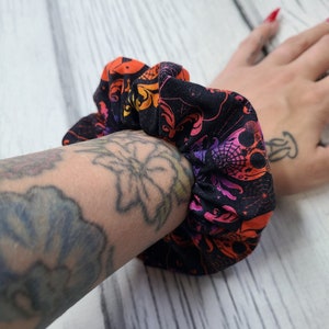 May include: A fabric hair scrunchie featuring a black background with a repeating pattern of skulls, spiderwebs, and ornate flourishes in vibrant shades of orange, pink, and purple. The accessory has a gathered, elasticized construction.