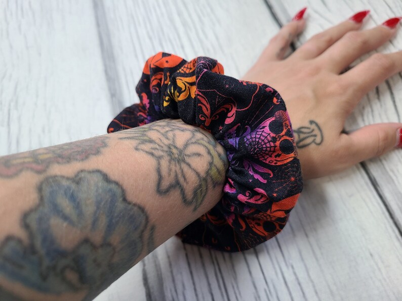May include: A fabric hair scrunchie featuring a black background with a repeating pattern of skulls, spiderwebs, and ornate flourishes in vibrant shades of orange, pink, and purple. The accessory has a gathered, elasticized construction.