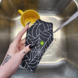 Pode incluir: Uma esponja de cozinha de tecido preto e branco com um desenho de teia de aranha e uma aranha verde no centro. A esponja está sendo enxaguada em uma pia de aço inoxidável.