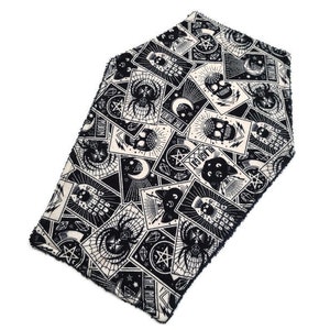 May include: A black and white coffin-shaped table runner with a repeating pattern of tarot card-style illustrations. The design includes skulls, spiders, moons, and pentagrams. The edges have a fringed trim.