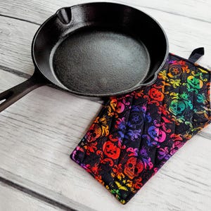 May include: A black cast iron skillet with a long handle rests next to a colorful, quilted pot holder. The pot holder features a black background with a repeating pattern of skulls, pumpkins, and other spooky designs in rainbow colors.