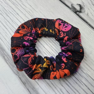 May include: A fabric hair scrunchie featuring a black background with a repeating pattern of orange and purple pumpkins, bats, and spider webs. The scrunchie has a gathered, elasticized design that creates a ruffled, circular appearance.