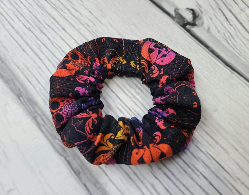 May include: A fabric hair scrunchie featuring a black background with a repeating pattern of orange and purple pumpkins, bats, and spider webs. The scrunchie has a gathered, elasticized design that creates a ruffled, circular appearance.